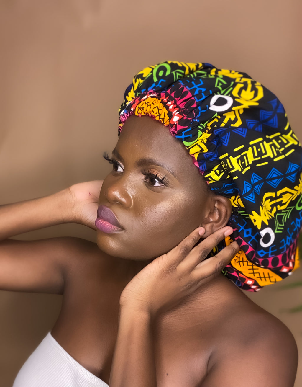 Brown skin model wearing a large satin lined bonnet in multicolor like yellow, pink, white, black, green and blue. This bonnet is a kente bonnet, african print satin lined, ankara bonnet, hair bonnet. Lined with Pink Satin
