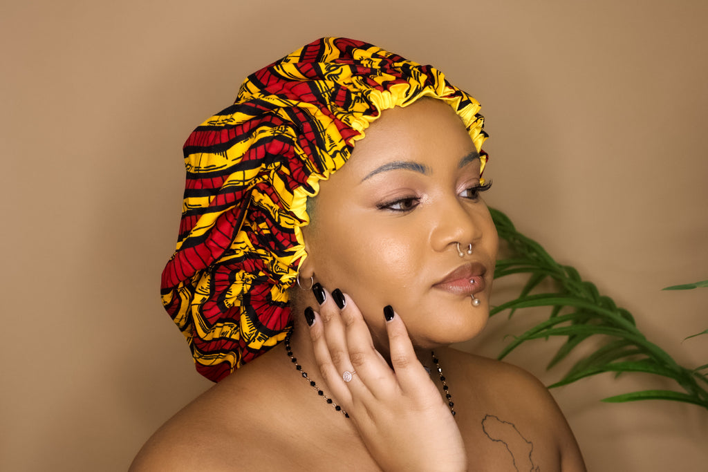 Brown skin model wearing a large satin lined bonnet in multicolor like red, yellow, white, black. This bonnet is a kente bonnet, african print satin lined, ankara bonnet, hair bonnet. Lined with yellow satin. 