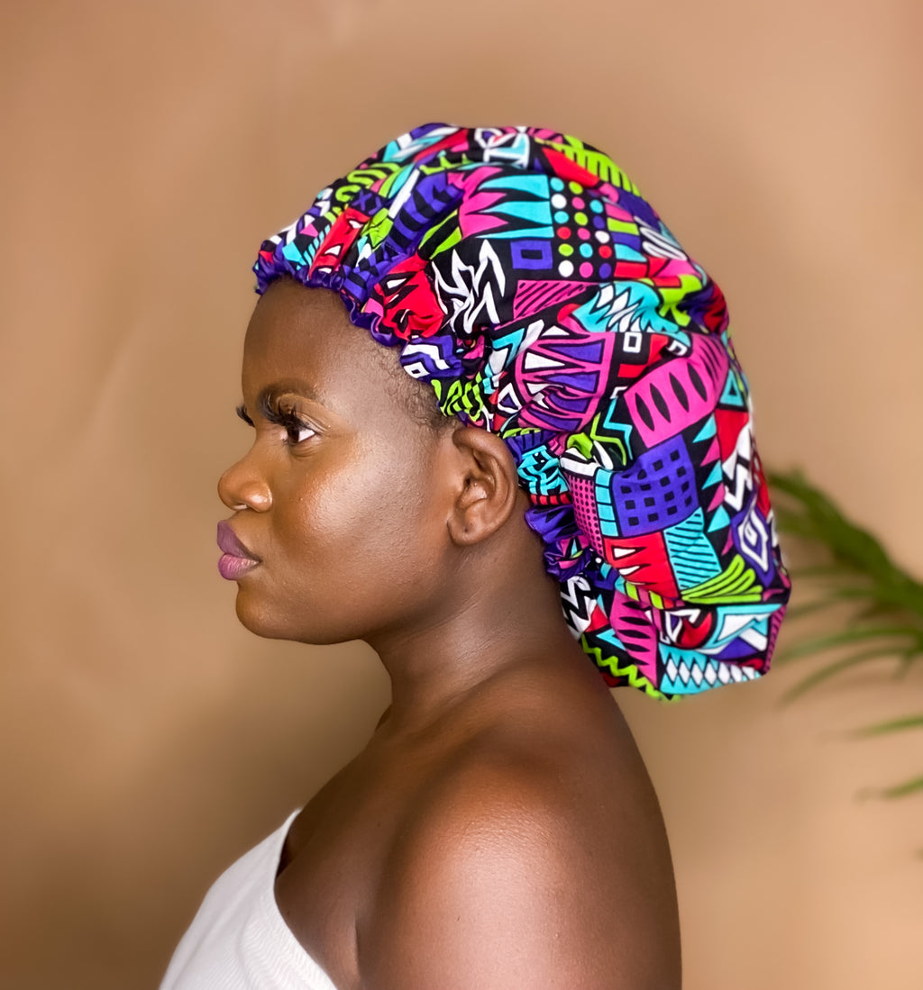 Brown skin model wearing a large satin lined bonnet in multicolor like purple, pink, white, black, green and blue. This bonnet is a kente bonnet, african print satin lined, ankara bonnet, hair bonnet